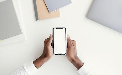 Руки дорослої людини тримають смартфон з порожнім білим екраном над столом, поруч лежать зошити та ноутбук.